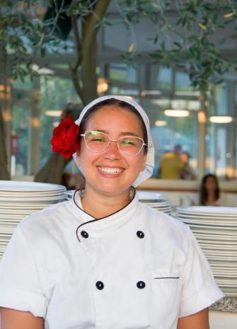 Chef souriante avec fleur rouge dans les cheveux, devant des assiettes empilées.