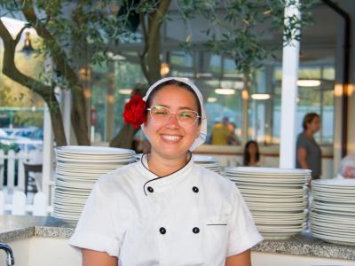 Chef souriante avec fleur rouge dans les cheveux, devant des assiettes empilées.