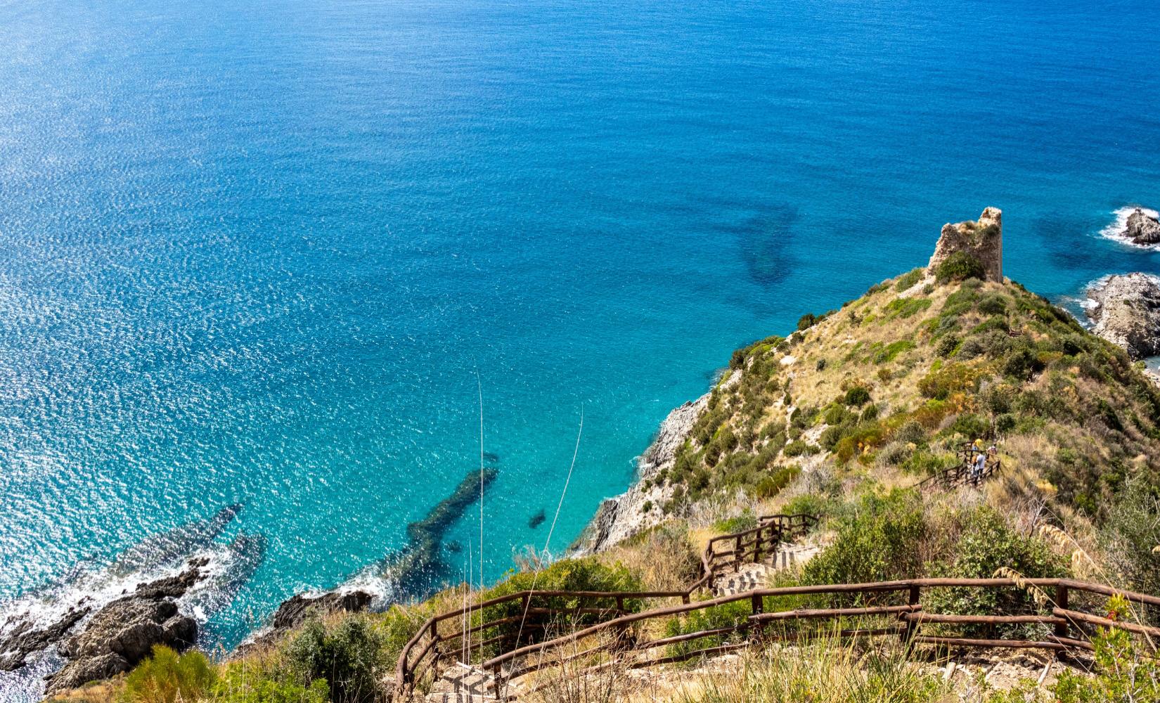 Scogliera con torre antica e mare cristallino, sentiero panoramico immerso nella natura.