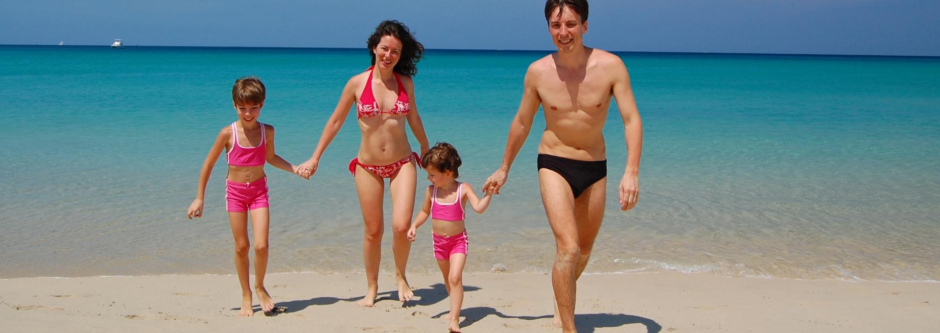 Famiglia felice cammina sulla spiaggia, mare turchese e cielo sereno sullo sfondo.