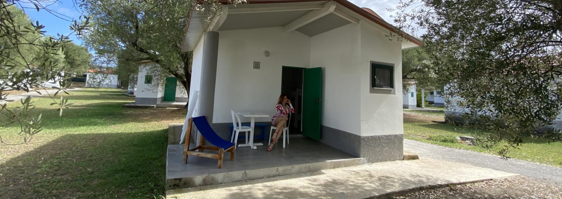 Bungalow bianco con veranda, sedia a sdraio e persona seduta all'aperto, circondato da alberi.