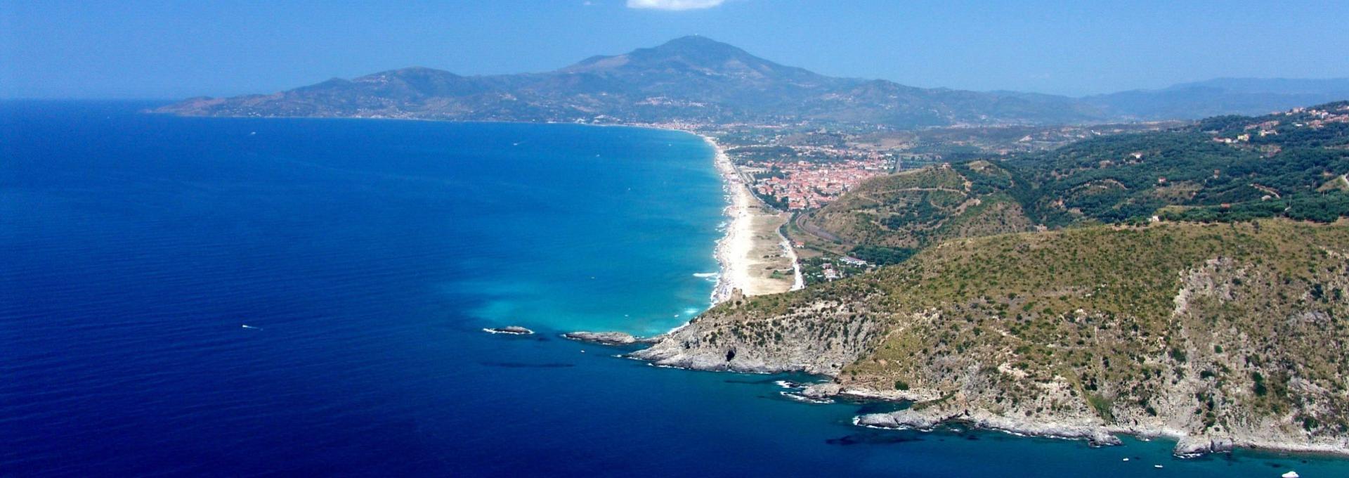 Costa rocciosa e mare blu con spiaggia sabbiosa e città in lontananza.