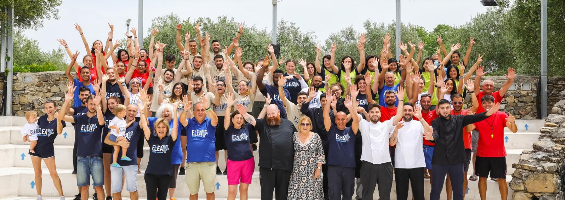 Gruppo di persone sorridenti all'aperto, indossano magliette colorate e sollevano le mani.