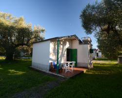 Piccola casa bianca con veranda, tavolo e sedie, circondata da alberi e prato.