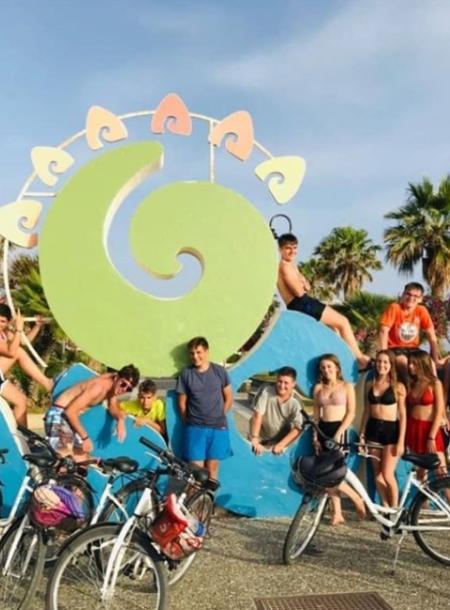 Gruppo di ragazzi in bici davanti a una scultura colorata con palme sullo sfondo.