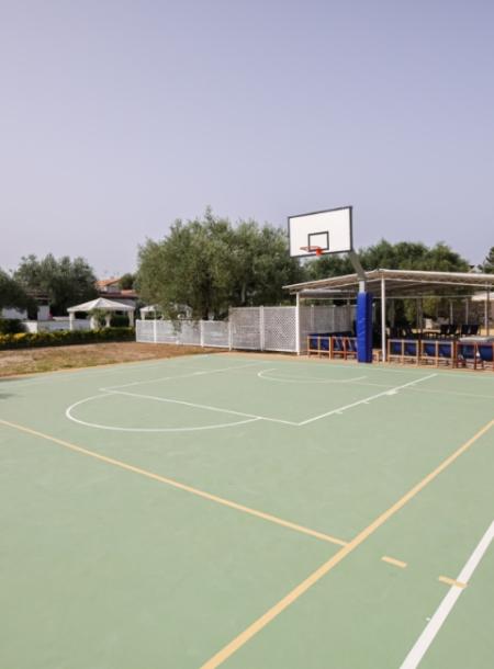 Campo da basket all'aperto con tabellone, circondato da alberi e panchine.