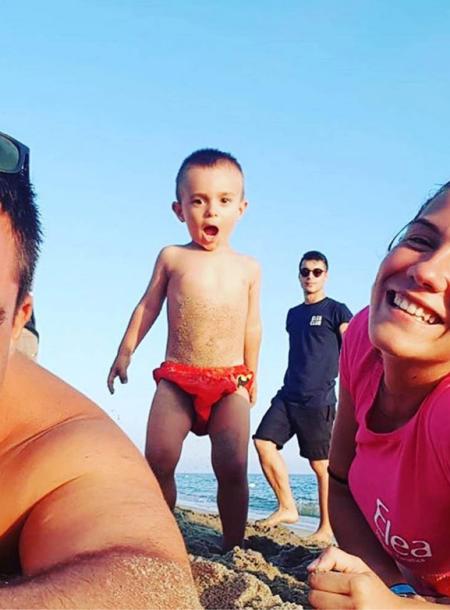 Famiglia in spiaggia, bambino sorpreso al centro, sorrisi e sole estivo.