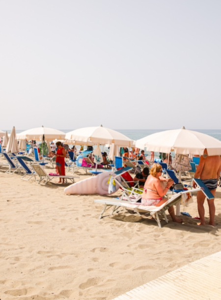 Spiaggia affollata con ombrelloni bianchi, lettini e bagnanti sotto il sole estivo.