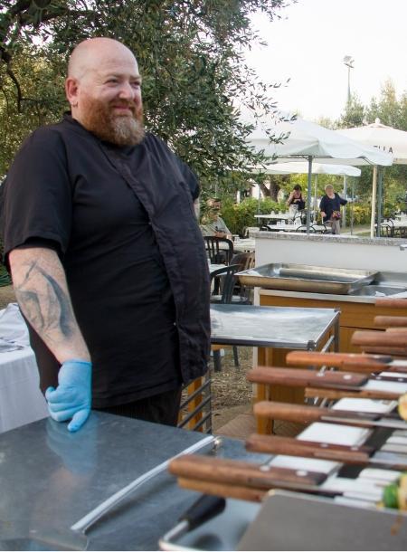 Chef con guanto blu, sorridente, accanto a tavolo all'aperto, atmosfera rilassata.