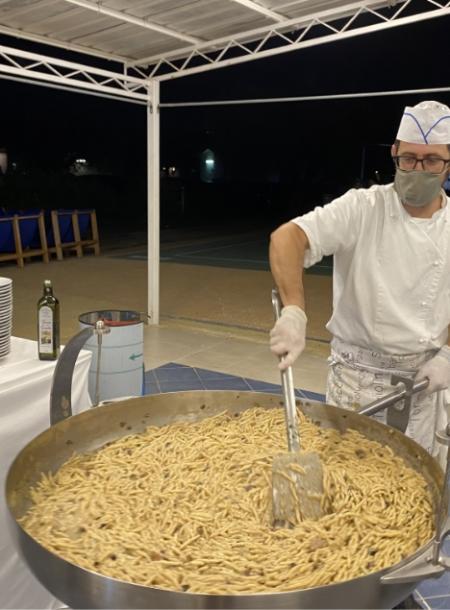 Cuoco prepara pasta in una grande padella all'aperto, con olio d'oliva a lato.