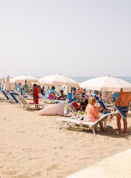 Spiaggia affollata con ombrelloni bianchi, lettini e persone che si rilassano al sole.