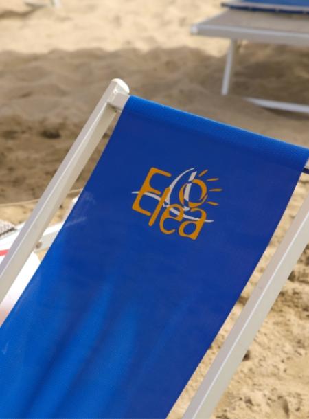 Sdraio blu sulla spiaggia con logo Elea in giallo e bianco.