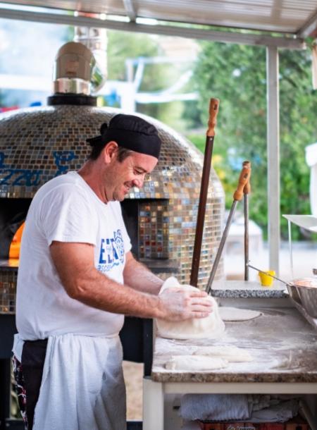 Pizzaiolo prepara l'impasto davanti a un forno a legna all'aperto.