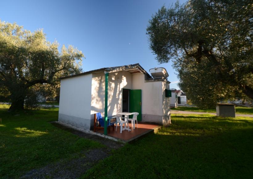 Piccola casa bianca con veranda, tavolo e sedie, circondata da alberi e prato.