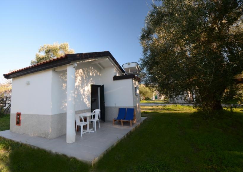 Piccola casa bianca con veranda, sedie bianche e sdraio blu in giardino verde.